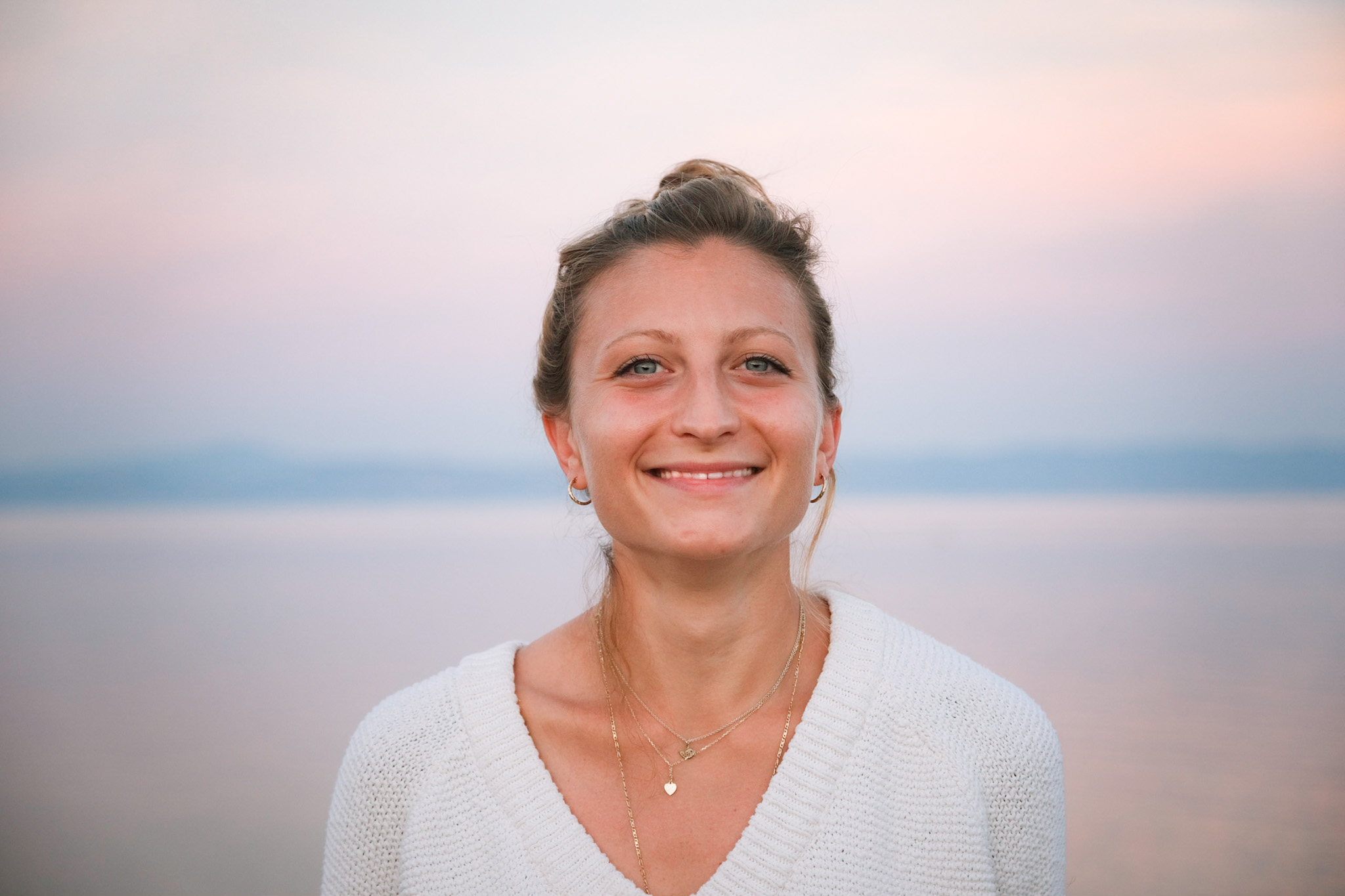 A woman smiles a the camera with a glowing landscape behind her and her blonde hair tied back