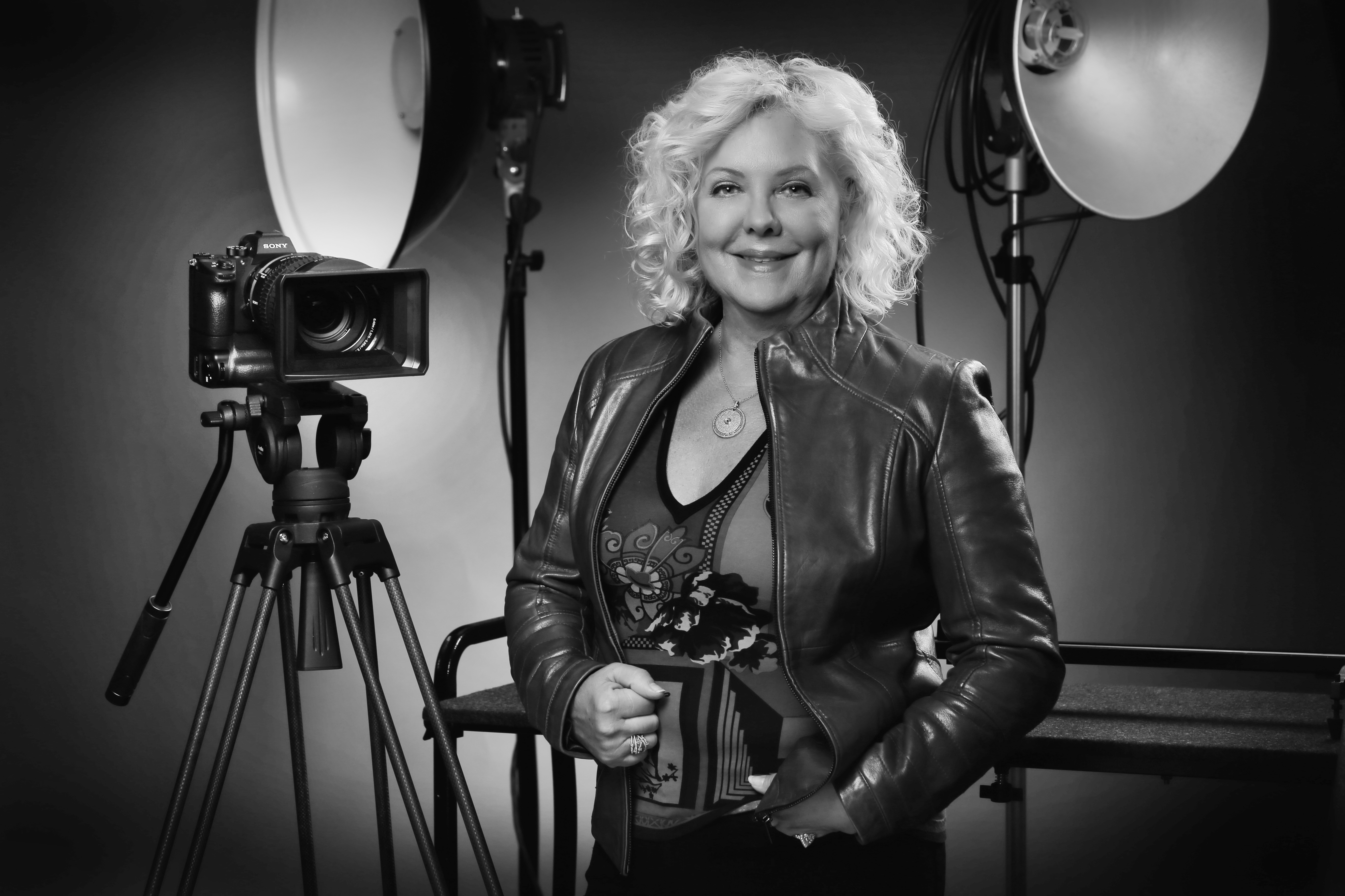 Photo of blonde woman standing by film camera with spotlights in background.