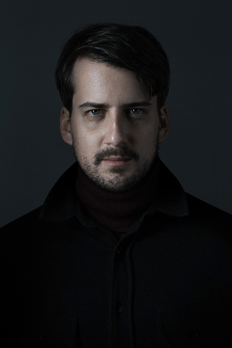 Image of a White man with mustache wearing dark shirt standing in front of dark background