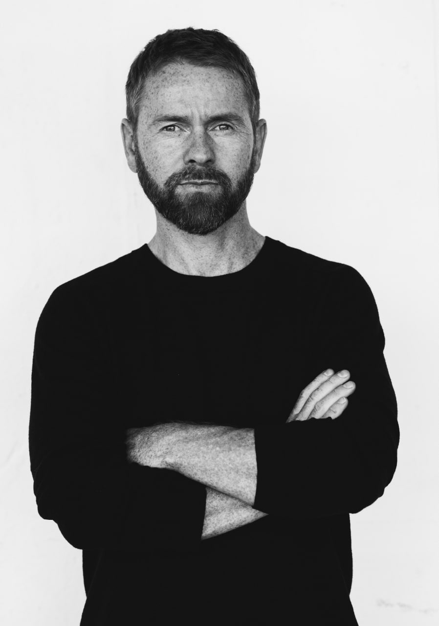 Black and white photo of man crossing his arms with dark hair and facial hair