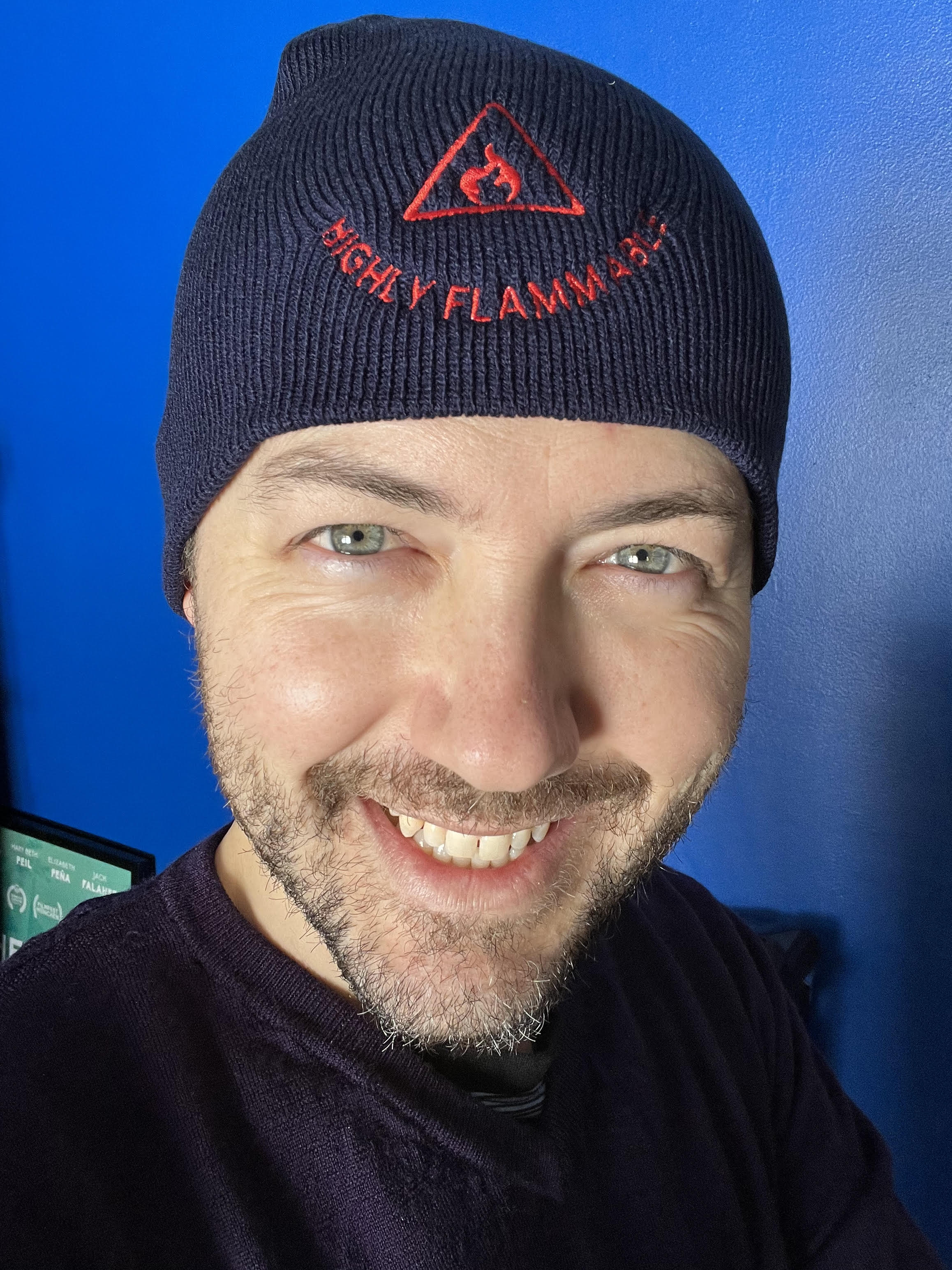 Image of White man smiling broadly at camera wearing a black hat on his head, and a black shirt in front of a blue background.