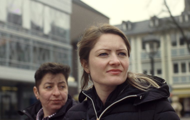 Photo of white woman with blonde hair looking out in distance. Behind her a woman with short hair looks over her shoulder as well.