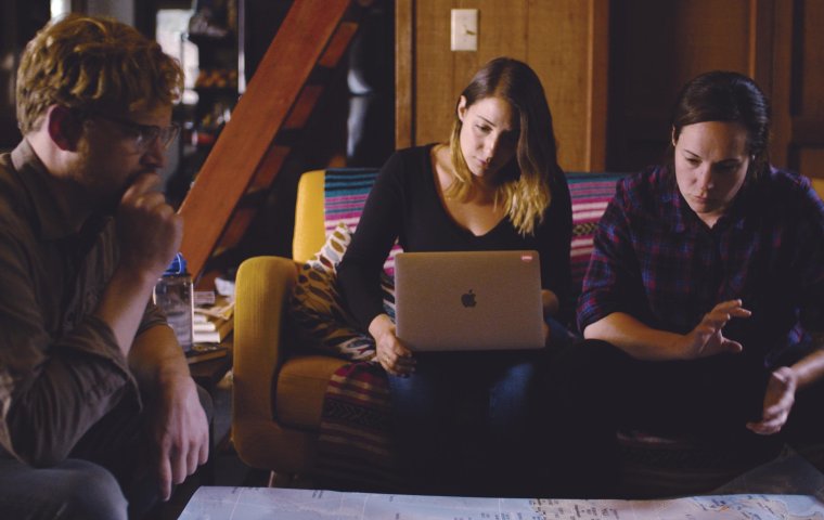 Three people sit around a low table with a large sheet of paper looking intently, while the central figure, a woman, looks down at her Mac Laptop 