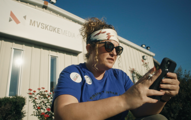 Image of Native American woman with brown hair in a ponytail, and sunglasses, looking at her phone. 