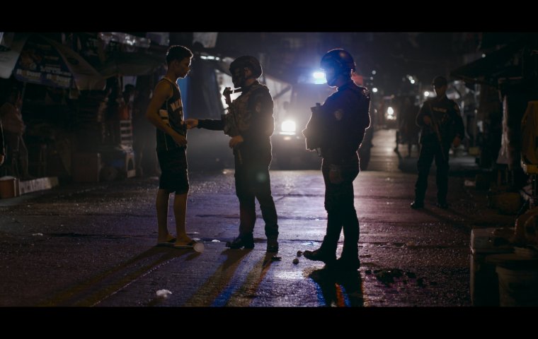 A shadowy image taken at night, backlight by a car's headlamps. Two men are in combat gear with guns and helmets speaking to a young person in tshirt and shorts.