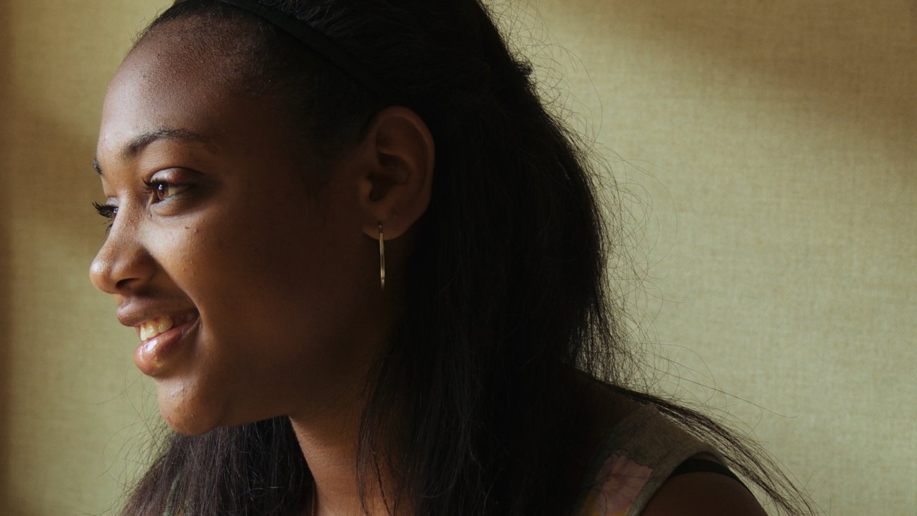 Photograph of a Black teenage girl with hair pulled back from her face, looking to the left smiling. 