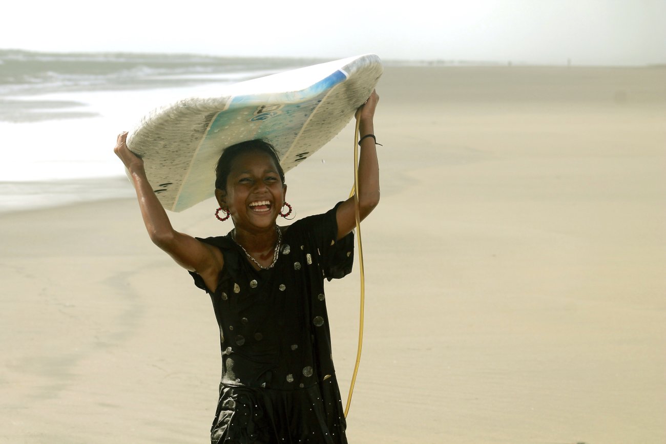 Young girl laughing as she holds a surf board on her head.