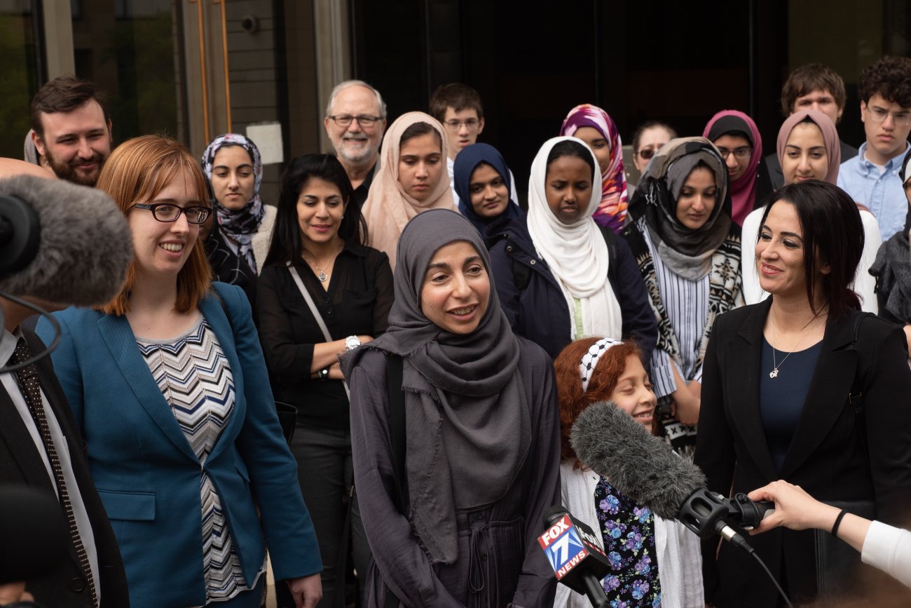 Photo of a crowd and a press conference, with a woman in front speaking with a Hijab.