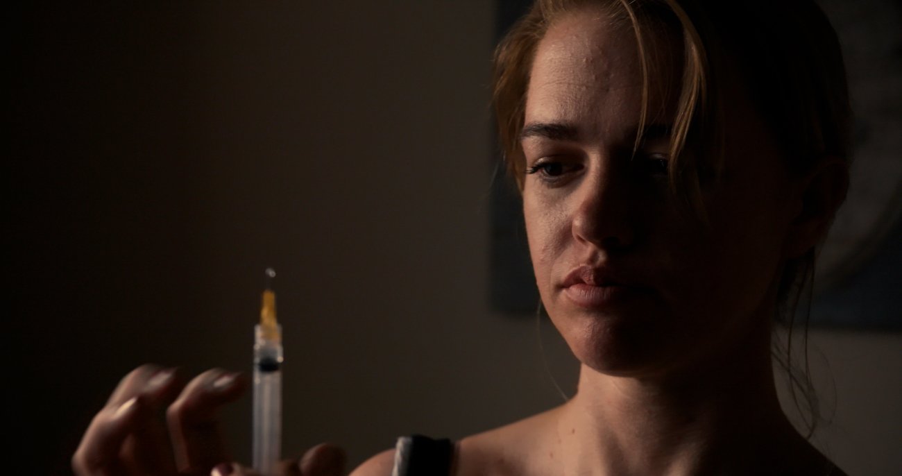 Image of a young white woman with brown hair pulled back to ponytail. She looks at a vial and stands in front of a dark background.