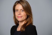 Image of White woman with light brown har wearing black shirt standing in front of gray background.