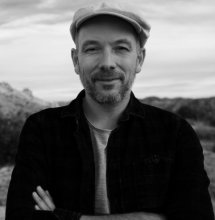 Black and white photo of man with a cap and facial hair folding arms